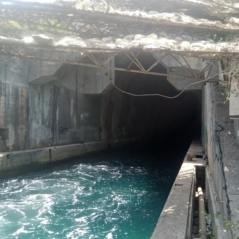 The dark arched mouth of a former submarine tunnel cut into the rock at the water's edge in the Bay of Kotor, the channel water glowing teal