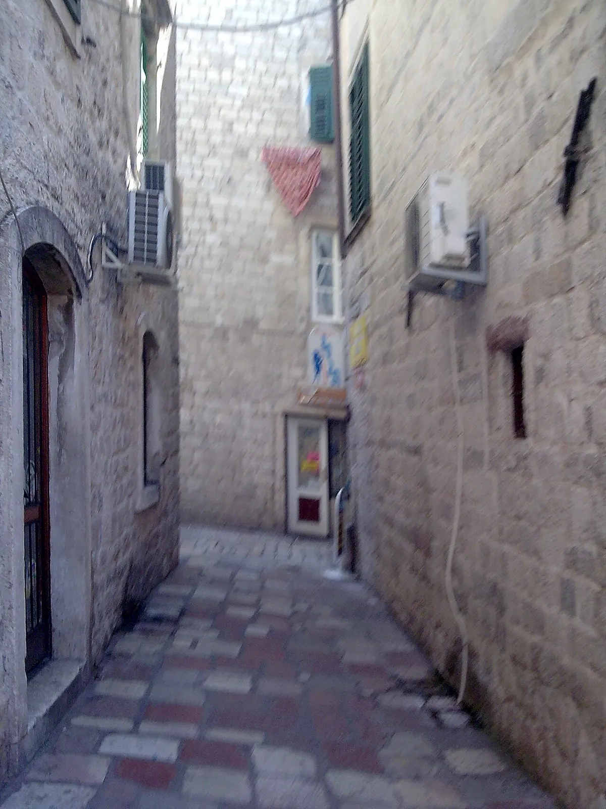 A narrow flat lane in Kotor paved with uneven stone slabs, running between tall historic stone buildings with shutters