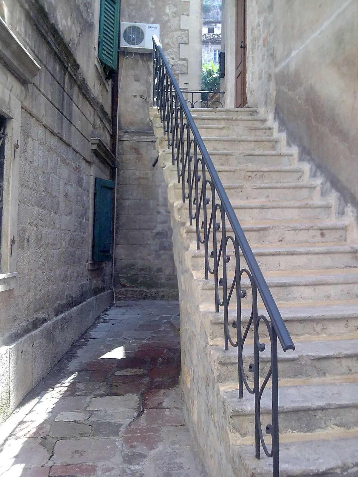 A narrow stone Old Town lane in Kotor with a steep stone stairway and wrought-iron handrail rising past apartment doorways