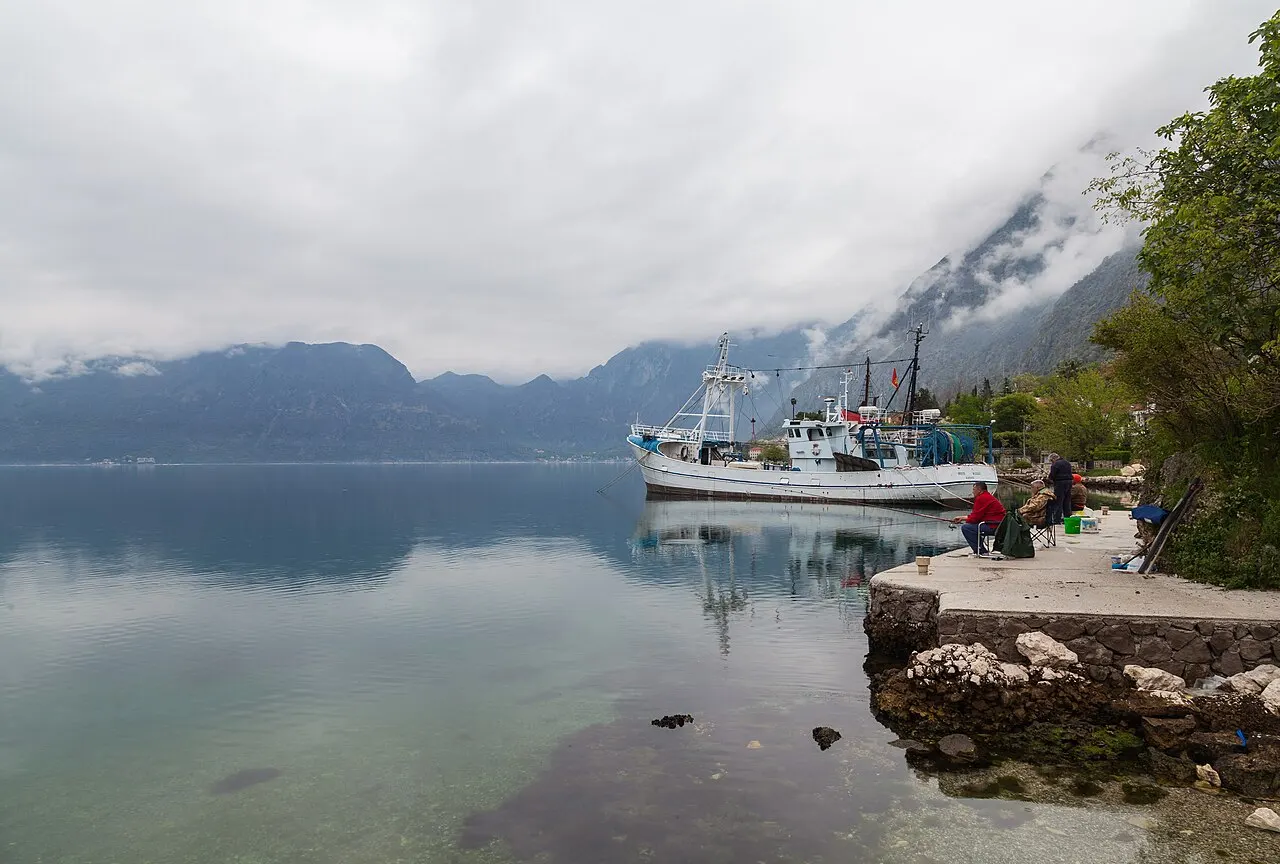 The calm Dobrota waterfront on the Bay of Kotor, a fishing boat moored at a stone quay with mist-wrapped mountains beyond