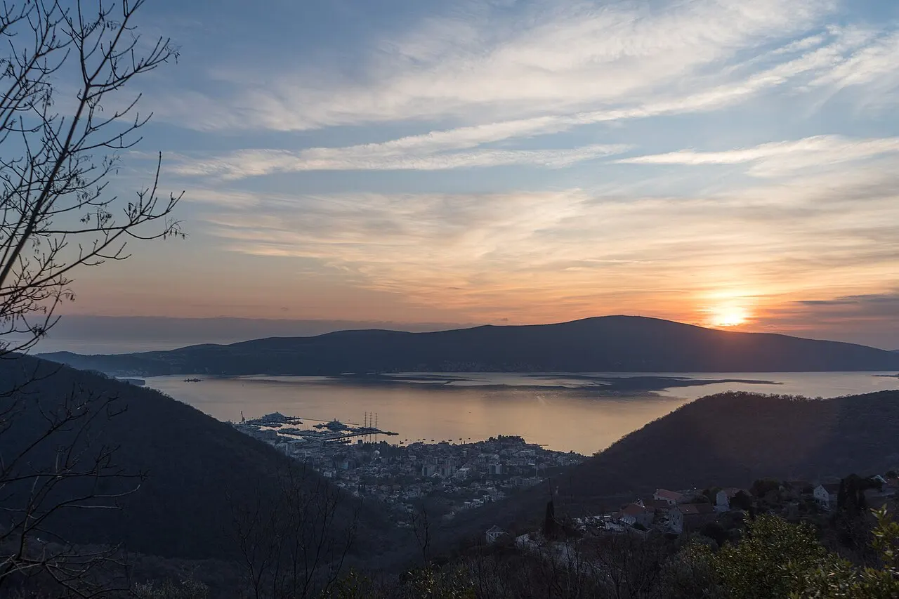 Golden-hour view from a high viewpoint over the Bay of Kotor, the town and waters far below as the sun sets behind the hills
