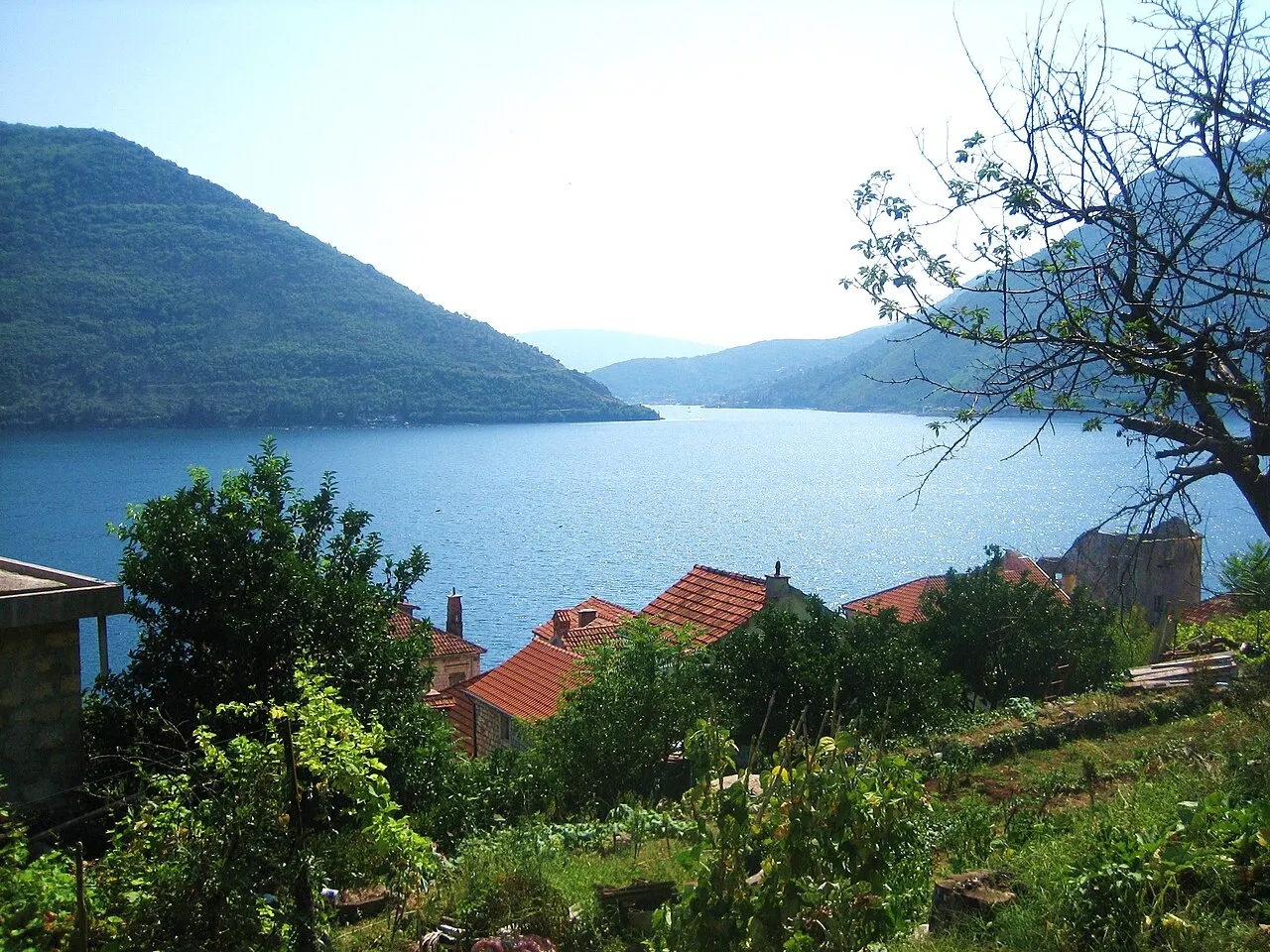 A sunlit view over Perast rooftops across the glittering Bay of Kotor toward green mountains and the narrows of the bay