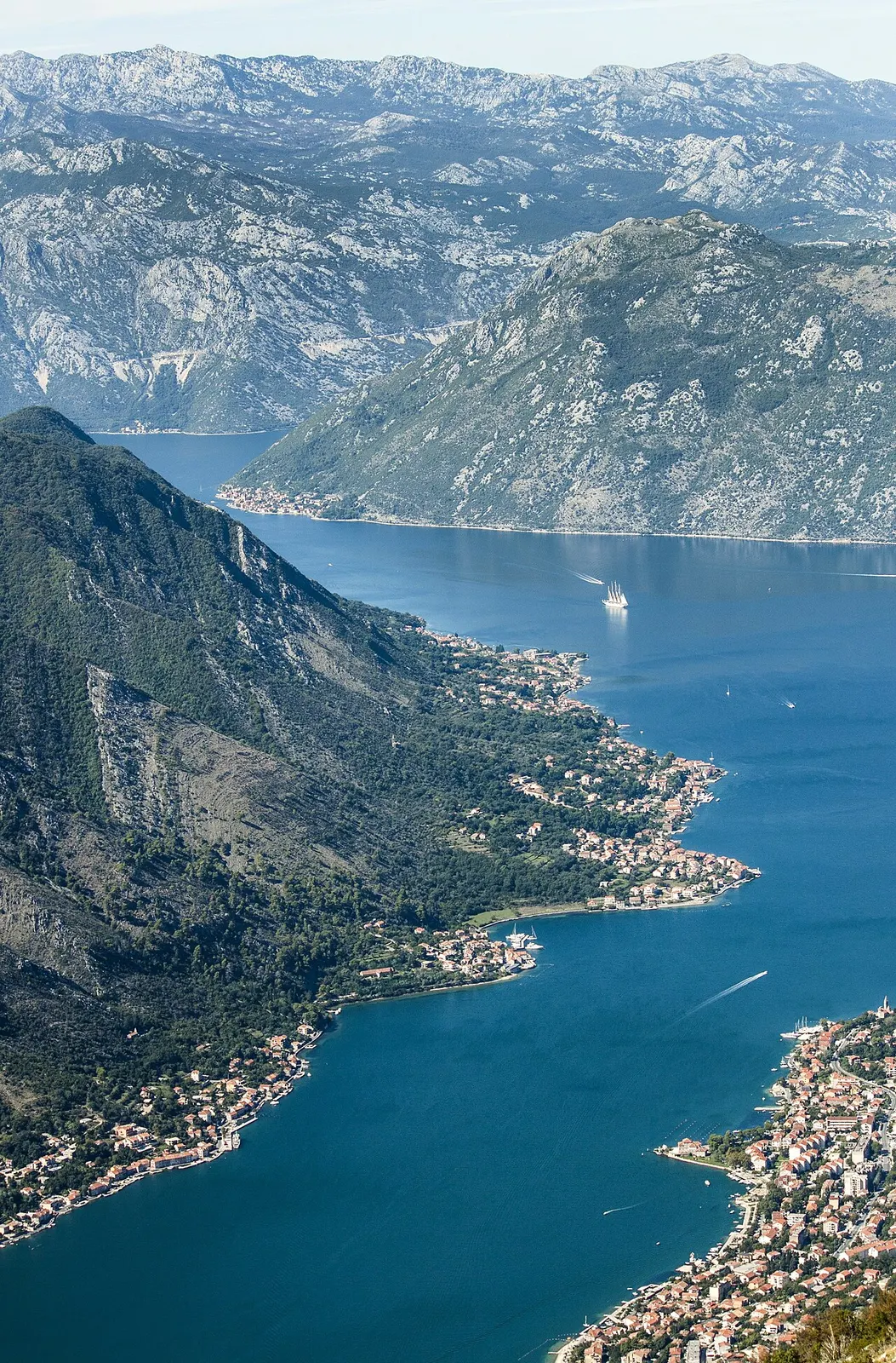 Deep-blue Bay of Kotor on a sunny day, small boats leaving wakes on the glassy water below steep wooded cliffs