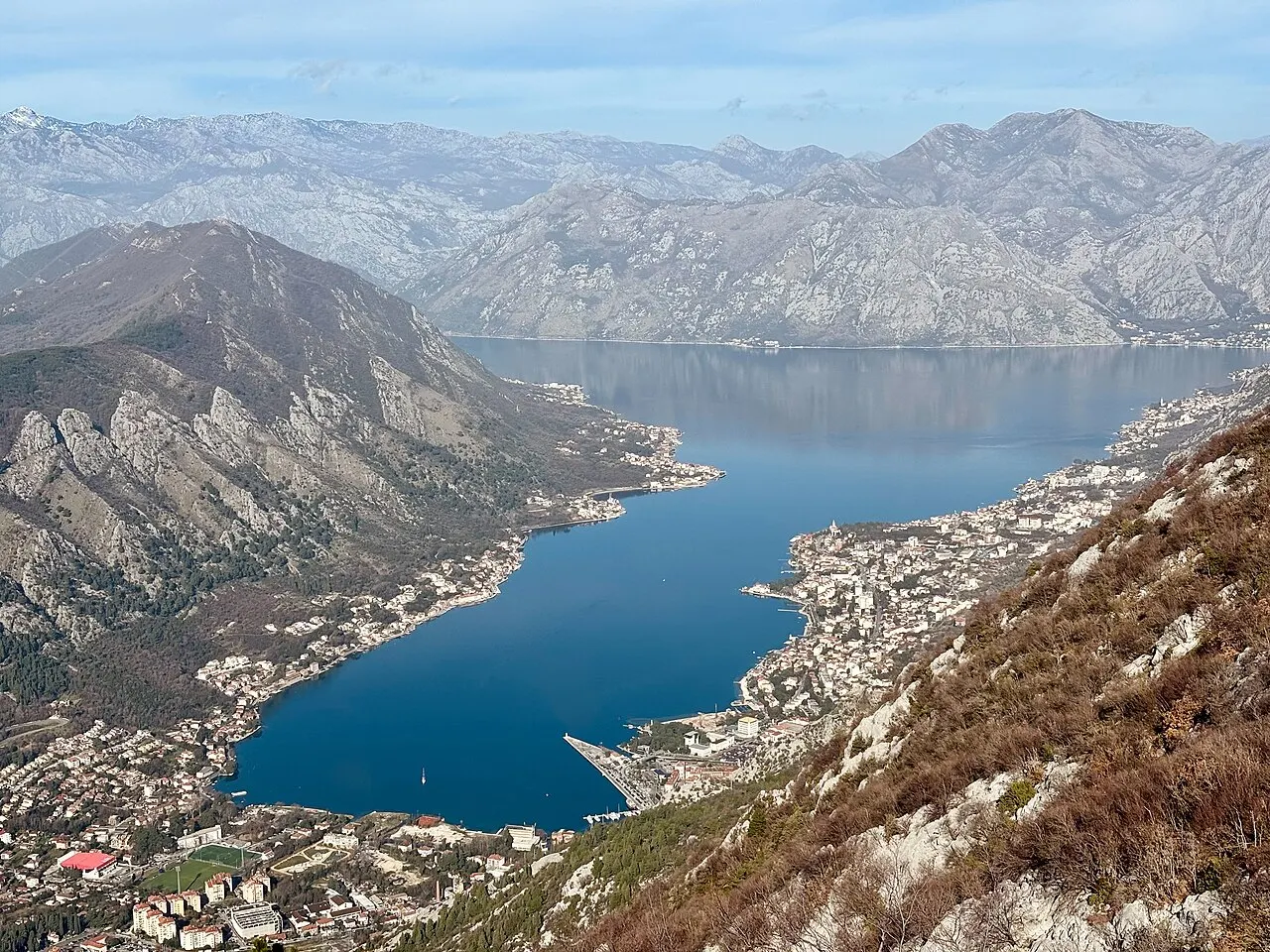 Winter view over Kotor and the quiet bay from above, bare slopes in muted light and snow on the distant peaks