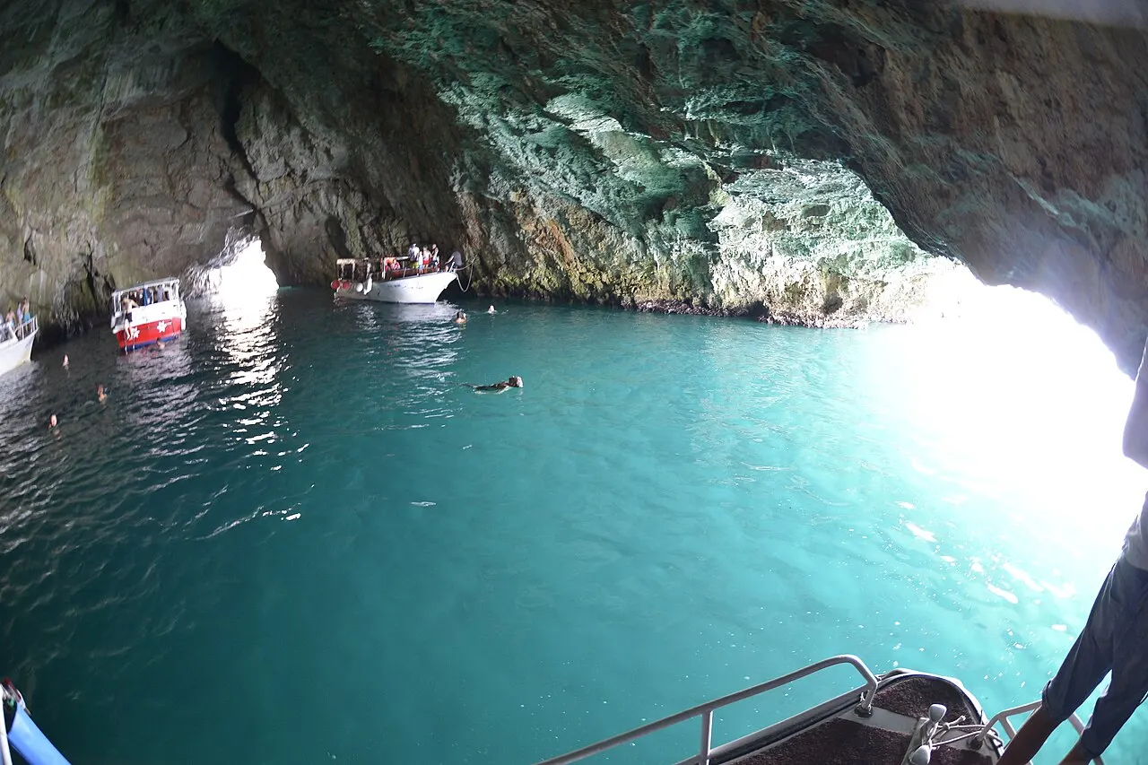 A swimmer floating in the glowing blue water inside the Blue Cave, tour boats moored by the wall and light at the mouth