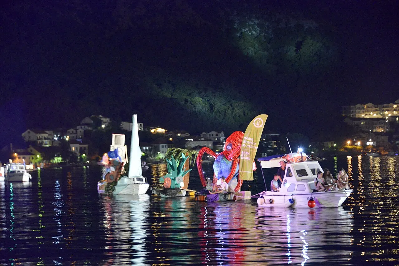 Decorated, brightly lit boats parading across the dark Bay of Kotor at night during Boka Night, shore lights beyond