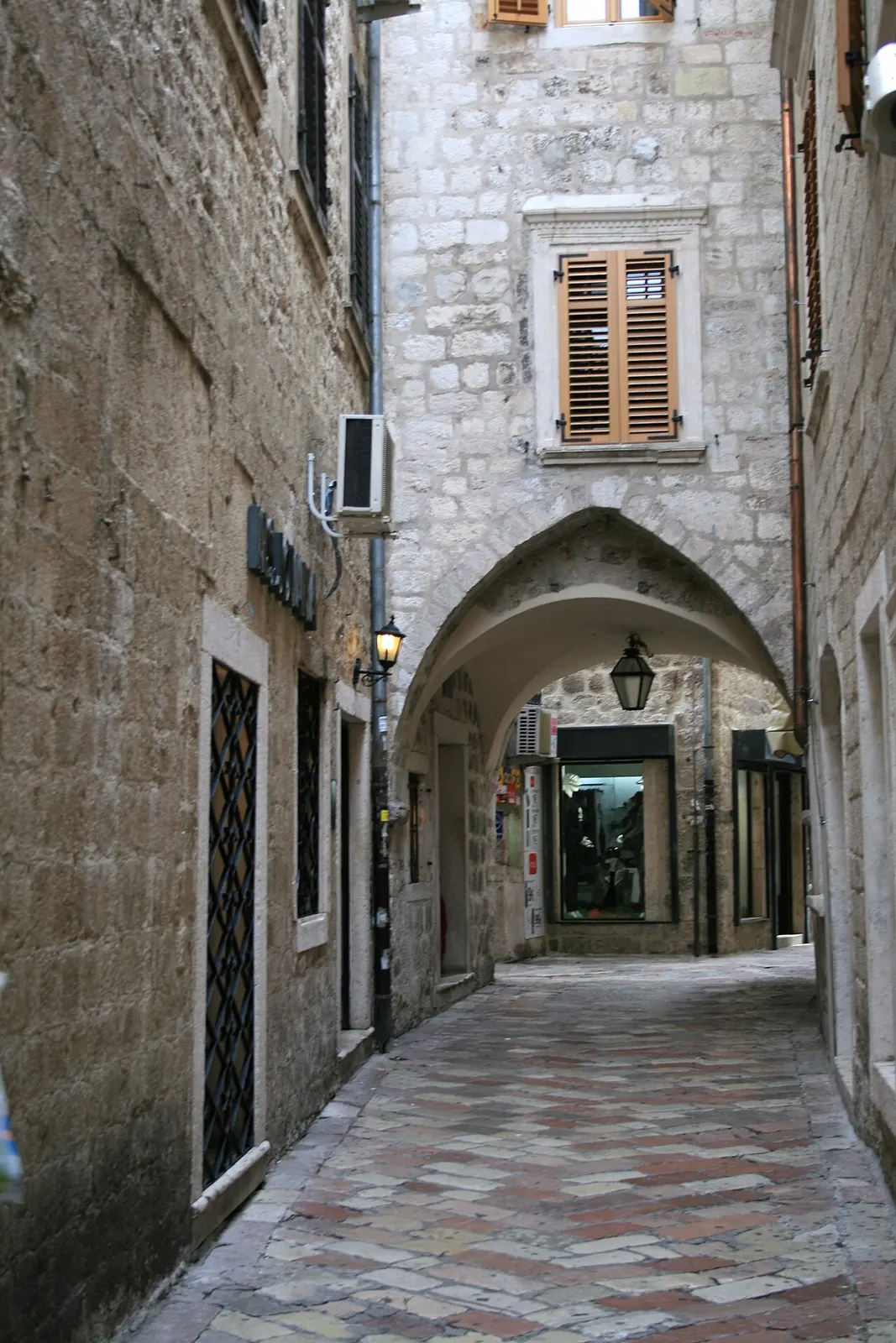 A quiet empty narrow stone street in Kotor's Old Town with a low archway, shuttered window and a wall lantern, no crowds