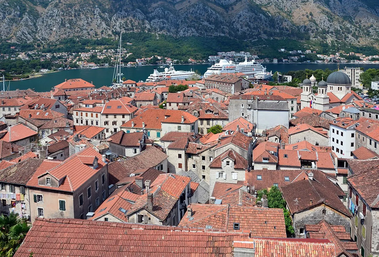 Kotor's terracotta Old Town rooftops seen from the city-wall path, the blue Bay of Kotor and a cruise ship beyond