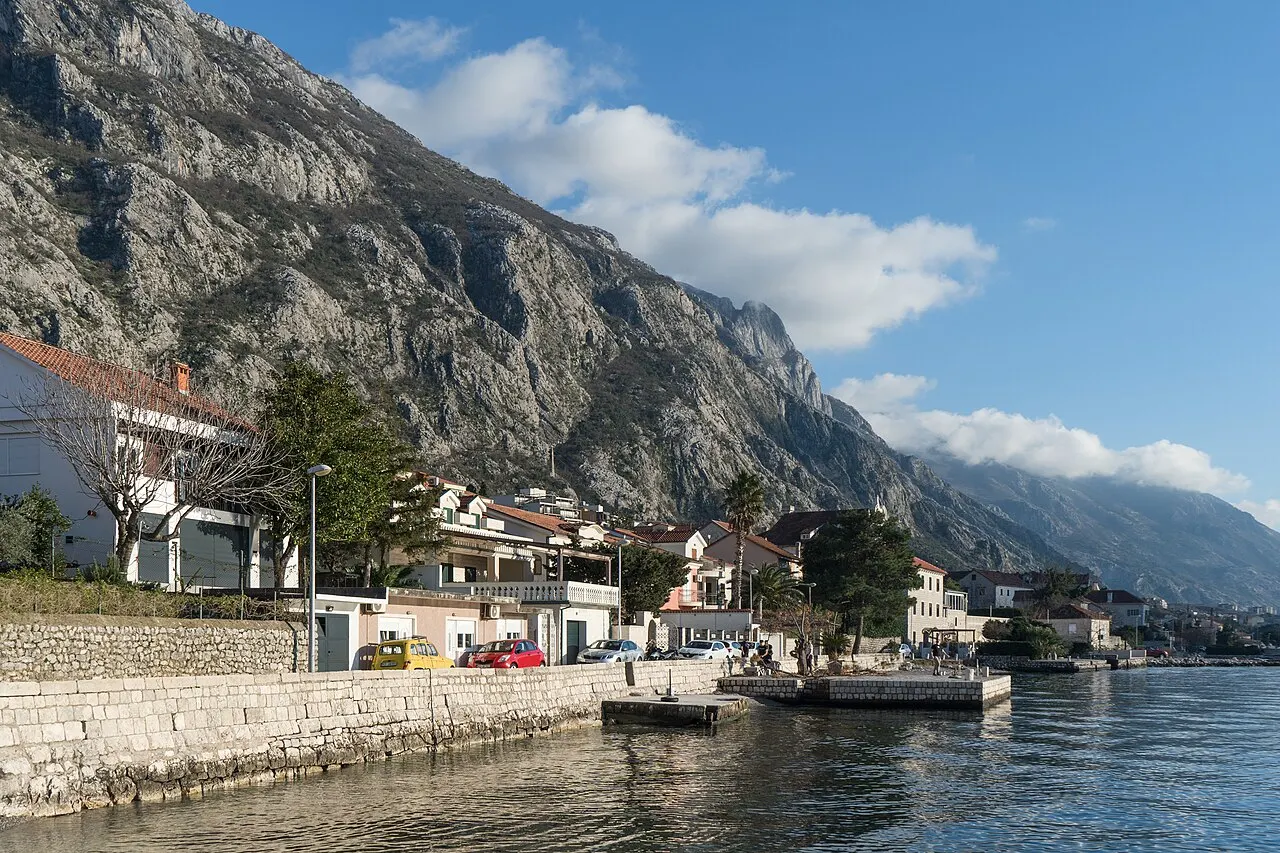 The Dobrota bayfront with stone houses, parked cars and a small quay, mountains rising across the Bay of Kotor
