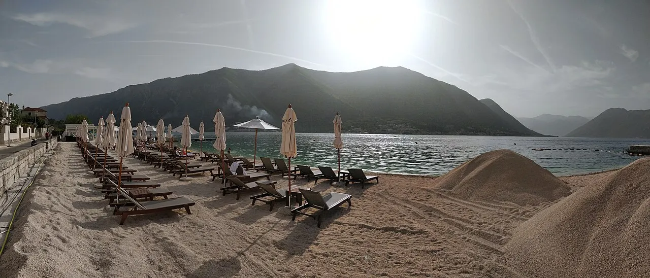 A Dobrota bay beach with sun loungers and umbrellas, the calm water and mountains beyond, a family swimming spot
