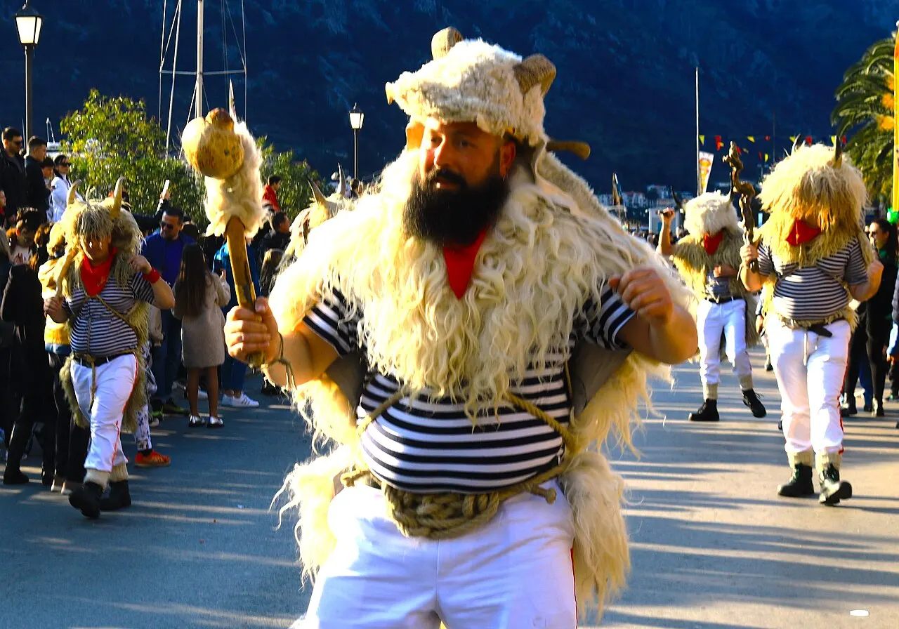 Costumed Carnival figures dancing through a Kotor street with crowds lining the waterfront
