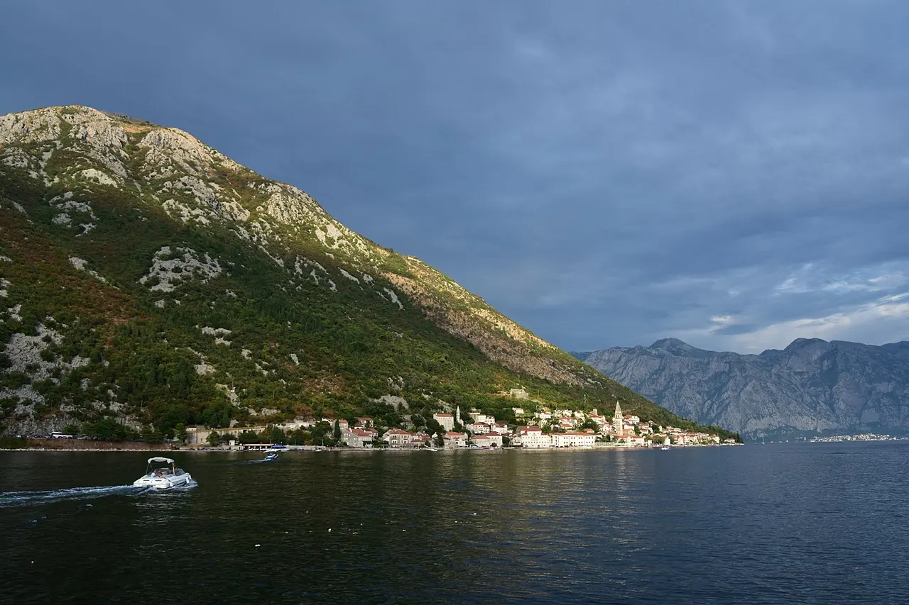 A small boat on the calm Bay of Kotor with Perast and its bell tower below dramatic mountains