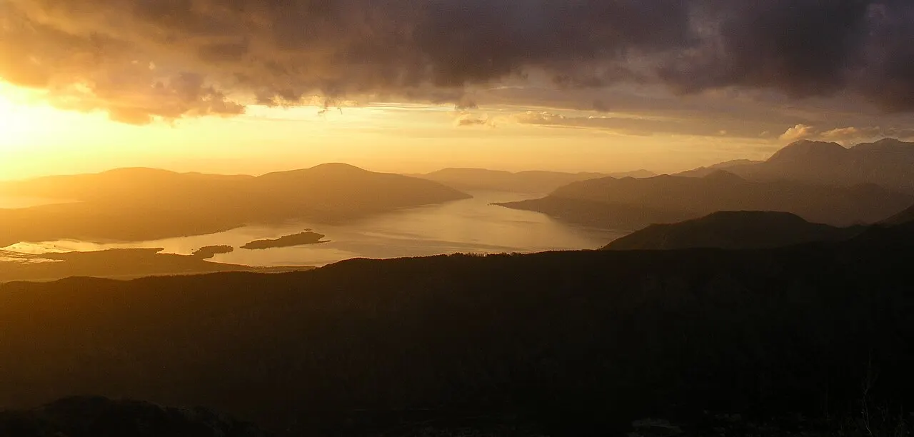 The calm Bay of Kotor at golden hour, the water glowing under a dramatic sunset sky