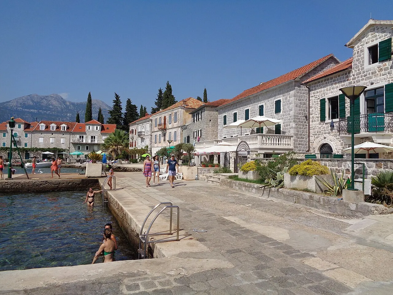 The quiet stone waterfront promenade of Rose village on Luštica, calm water and mountains across the bay